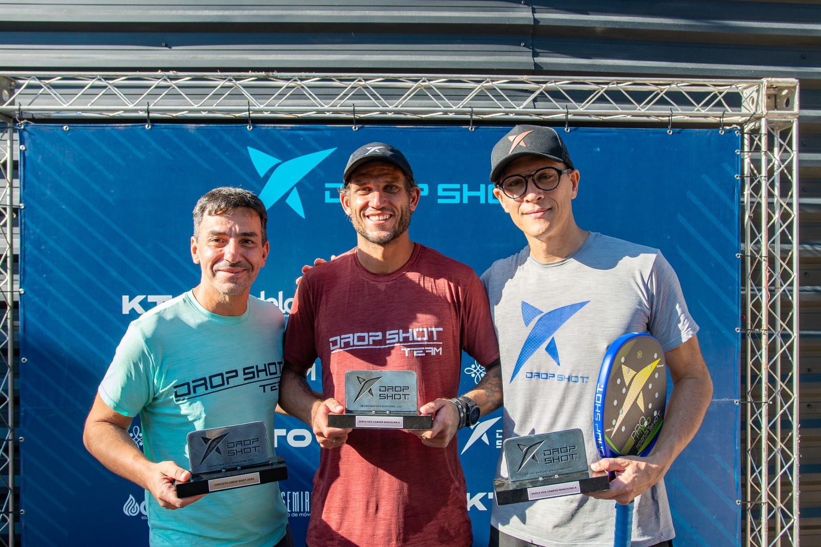Rafael Moura, de vermelho, ao centro com o troféu do Beach Tennis com Pablo Oliveira à direita, de cinza. À esquerda, Ricardo Aloise / Crédito: Diogo Keng